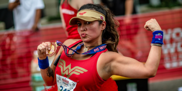 A women in Wonder Woman racing gear shows off her medal and flexes her arm muscles at the DC Super Hero Race