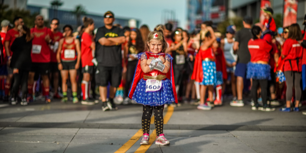 A young girl dressed up in a Wonder Woman costume poses in the crossed arm Wonder Woman pose while a crowd of racers stands behind her at the DC Super Hero Run