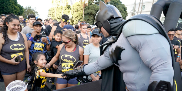 A photo of Batman reaching his arm out to a young girl in a crowd of people watching the DC Super Hero Run. Everyone is wearing Batman themed clothing and accessories.